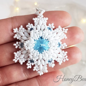 May include: A white beaded snowflake ornament with a light blue crystal in the centre. The snowflake is held in a hand against a blurred background.