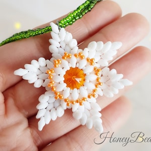 May include: A white beaded flower pendant with an orange crystal center, hanging from a green beaded chain. The pendant is shaped like a star and has a delicate, intricate design.
