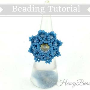 以下が含まれることがあります： 雪の結晶のデザインの青いビーズのリング。リングは白いディスプレイスタンドの上に置かれています。画像の右下隅に「Honey Beads1」というテキストがあります。