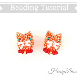 以下が含まれることがあります： 赤いビーズの襟が付いたオレンジ色のビーズでできた猫のイヤリングが2つ。猫は白い顔と緑色の目をしています。イヤリングは白い背景の上にあります。