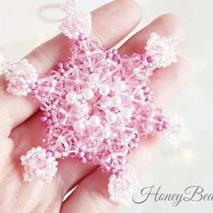 May include: A pink and white beaded snowflake ornament with a six-pointed star shape. The snowflake is made with small glass beads and pearl beads.