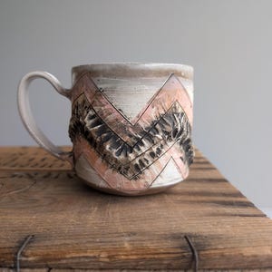 May include: A handmade ceramic mug with a cream-colored handle and rim. The mug features a chevron design in pink and black, with a textured, raised pattern. The mug is sitting on a wooden surface.