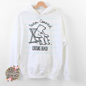 May include: White hoodie with a front graphic. The graphic features a cartoon bear in a beach chair with the text "Team Conrad" and "Cousins Beach" in black. The hoodie has a front pocket and a drawstring hood.