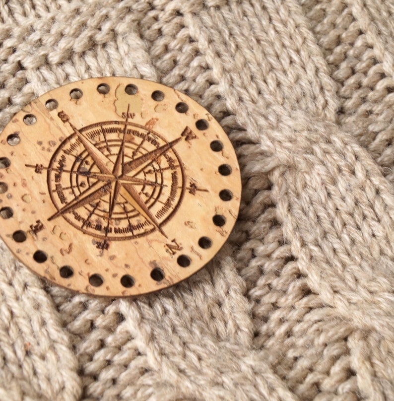 May include: A wooden compass with a brown background and a white compass rose design. The compass is surrounded by a ring of holes. The compass is on a beige knitted fabric.