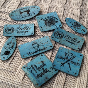 Könnte beinhalten: Sortiment an blauen Korkstoff-Etiketten mit verschiedenen Designs und Texten, darunter "Taylor's Attic", "Handmade", "Stella's Designs", "Hand Knitted", "Cali's Attic" und "Hand Made".
