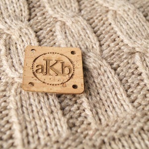 May include: A close-up of a brown cork label with the letters "akb" in a circle. The label is attached to a beige knitted fabric.