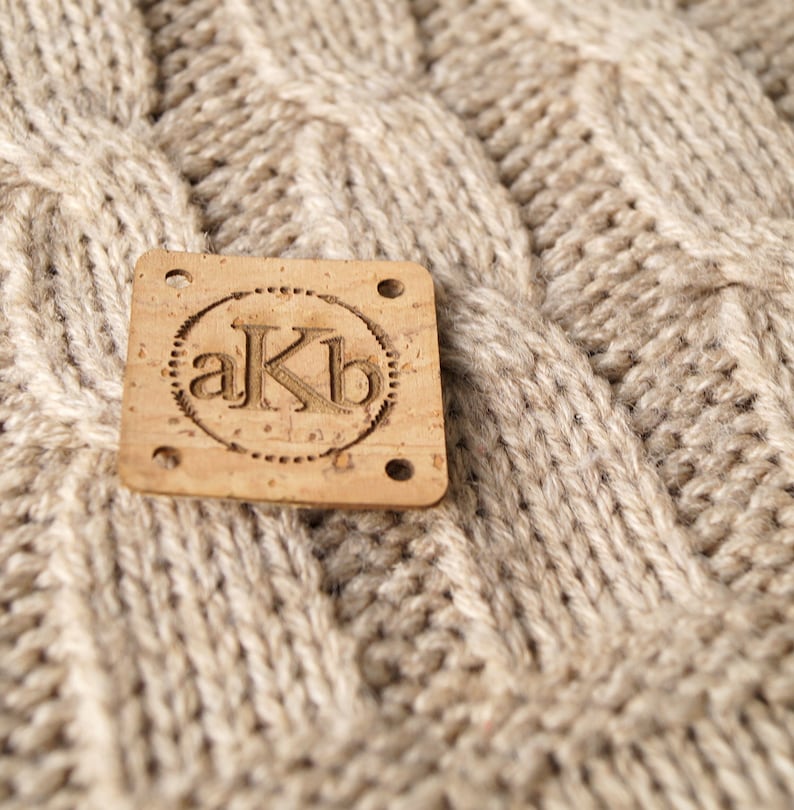 May include: A close-up of a brown cork label with the letters "akb" in a circle. The label is attached to a beige knitted fabric.