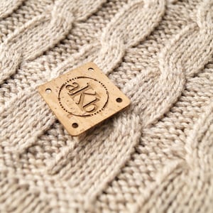 May include: A close-up of a cork label with the text "akb" in a circle. The label is on a beige knitted fabric.