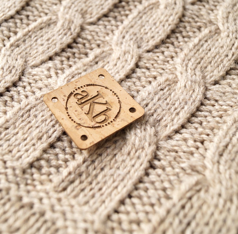 May include: A close-up of a cork label with the text "akb" in a circle. The label is on a beige knitted fabric.