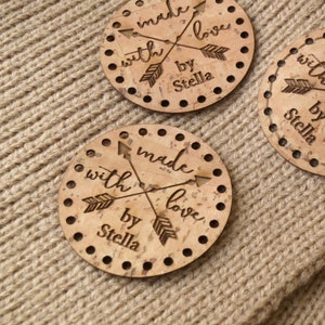 May include: Three round cork coasters with the text 'made with love by Stella' and two arrows crossing each other. The coasters are on a brown knitted surface.