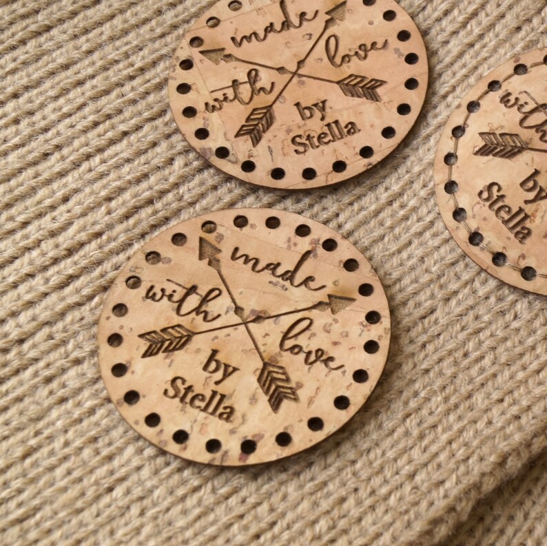 May include: Three round cork coasters with the text 'made with love by Stella' and two arrows crossing each other. The coasters are on a brown knitted surface.