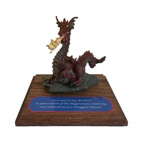 May include: A red dragon figurine with yellow flames coming out of its mouth, sitting on a black slate base. The figurine is on a wooden base with a blue plaque that reads "Presented to Jay Randall in appreciation of his imagination, creativity, and enthusiasm as a Dungeon Master."