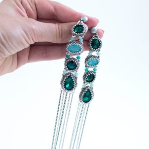 May include: Long, dangling earrings featuring emerald green and light blue gemstones, accented with silver-toned beads and chains. The earrings are held against a white background, showcasing their intricate design and color.
