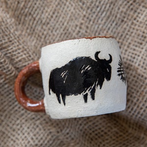 Pode incluir: Uma caneca de cerâmica branca com uma silhueta preta de um bisão pintada no lado. A caneca tem uma alça marrom.