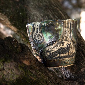 Könnte beinhalten: Ein handgefertigtes Keramikgefäß mit dunkler, strukturierter Außenseite. Das Gefäß zeigt eine schwarze Illustration eines Fuchses und einer Schlange. Die Farbpalette umfasst Grüntöne, Braun und Schwarz. Das Gefäß ruht auf einem Ast.