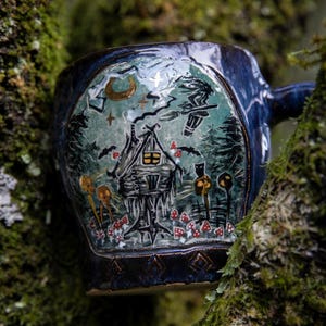 May include: Dark blue ceramic mug with a whimsical forest scene. The design features a cabin, bats, mushrooms, and a witch on a broomstick. The mug has a handle and is set against a mossy tree.