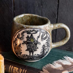 May include: A handmade ceramic mug with a whimsical illustration of a house in a forest, with a yellow window. The mug has a speckled, earthy green and brown glaze. Two lit candles and a feather are next to the mug.