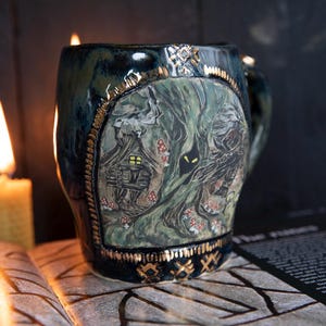 May include: A ceramic mug with a forest scene featuring a small house, trees, and mushrooms. The mug has a dark blue and green glaze with gold accents. A lit candle and an open book are in the background.