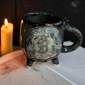 May include: A dark blue ceramic mug with a forest scene illustration, featuring a cabin, trees, and a crescent moon. The mug has a unique handle and three small feet. Two lit candles are in the background.