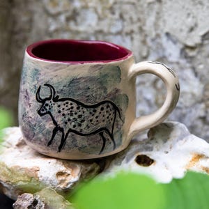 Puede incluir: Una taza de cerámica con exterior crema e interior burdeos. La taza presenta una ilustración negra de un toro manchado. El asa es de color crema. La taza está sobre una roca.
