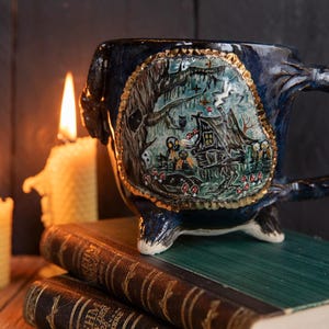 May include: A dark blue ceramic mug with a whimsical forest scene, featuring a house, trees, and figures, set against a gold trim. The mug sits on a stack of books, with lit candles in the background, creating a cozy, magical atmosphere.