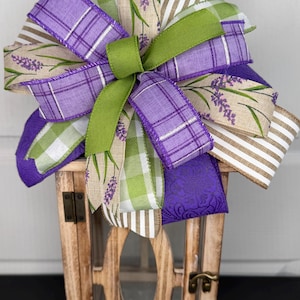 May include: A wooden lantern with glass panels, topped with a decorative bow. The bow features ribbons in purple plaid, green gingham, lavender sprig print, and tan and white stripes. The lantern has a rustic, farmhouse style.