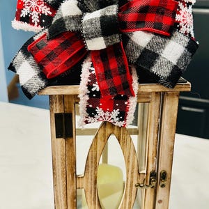 May include: A wooden lantern with a decorative bow. The bow features black and white buffalo plaid and red plaid ribbon with snowflake accents. The lantern has a glass panel and a candle inside.