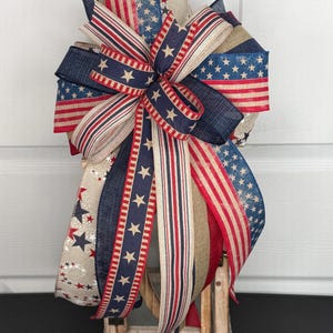 May include: A decorative bow crafted from multiple ribbons featuring red, white, and blue color schemes. The ribbons display patterns of stars, stripes, and solid navy blue textures, arranged in a multi-layered design with long, flowing tails.