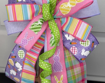 May include: A decorative Easter bow with multiple ribbons in various colors. The ribbons feature bunny and carrot patterns, stripes, and polka dots. The bow is accented with a green polka dot ribbon.