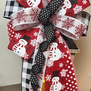 May include: A decorative Christmas bow featuring a snowman and snowflake design. The bow has red, white, and black ribbons with polka dots, snowflakes, and a snowman print. The bow is attached to a wooden lantern.