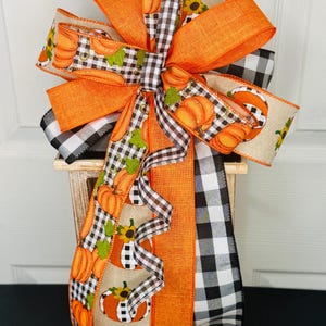 May include: A decorative bow crafted from multiple ribbons featuring orange solid fabric, black and white buffalo check patterns, and a tan burlap-style ribbon printed with orange pumpkins, green leaves, and yellow sunflowers, arranged in a large cluster.