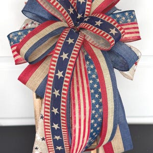 May include: A patriotic bow featuring red, white, and blue ribbons with star and stripe patterns. The bow is made of various textures and is likely used for decorative purposes. The bow is placed on a wooden lantern.