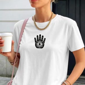 May include: A white short-sleeved t-shirt featuring a black graphic print of a hamsa hand with an eye in the centre. The shirt is paired with a thick gold-toned chain necklace and a pink shoulder strap visible on the side.