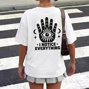 May include: A white short-sleeve t-shirt featuring a black graphic on the back. The graphic displays a stylized hand with an eye in the center, flanked by crescent moons and stars, with the text I NOTICE EVERYTHING printed below the design.