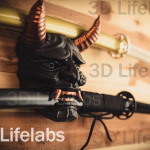 Pode incluir: Uma máscara Oni preta e cobre com chifres montada em uma parede de madeira com uma katana preta com um envoltório de corda preto e branco. O texto "3D Lifelabs" é visível na parede.