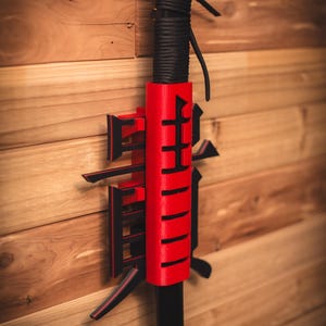 May include: A red and black sword holder with the Japanese word "武士" (Bushi) meaning "warrior" on the front. The sword holder is mounted on a wooden wall.