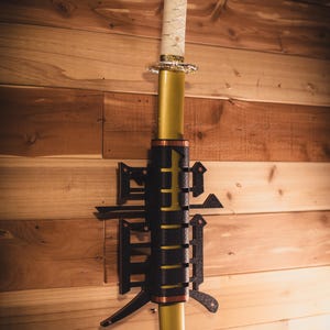May include: A gold-colored katana sword with a white and gold handle is mounted on a wooden wall with a black metal sword holder. The holder is shaped like a stylized Japanese character.