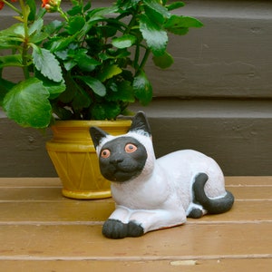 May include: A white ceramic cat figurine with black ears, eyes, and tail, lying on a wooden surface. A potted plant with green leaves and red flowers is in the background.