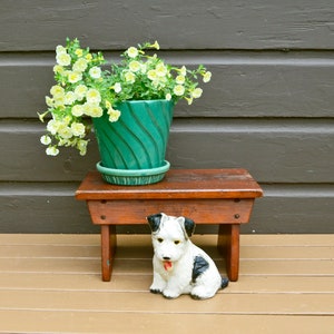May include: A small wooden stool with a white and black ceramic dog figurine sitting on top. A green planter with white flowers sits on the stool.