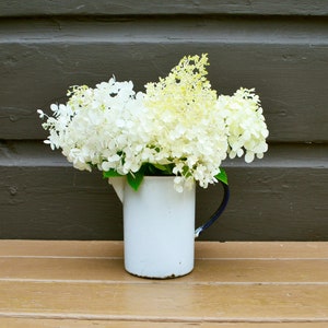 May include: A white enamel pitcher with a black handle holds a bouquet of white and light green hydrangea flowers. The pitcher sits on a wooden surface against a dark brown wooden background. The flowers are in full bloom, creating a soft, natural aesthetic.
