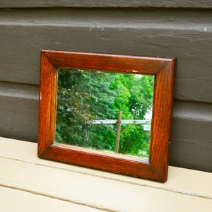 May include: A square, wooden-framed mirror with a rich, reddish-brown finish. The mirror reflects a vibrant outdoor scene with lush green trees and a glimpse of a fence. The mirror is resting on a light-colored surface.