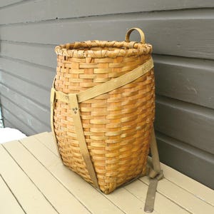 May include: A woven, light brown basket with a rectangular base and a rounded top. It features a handle and tan straps for carrying. The basket is made of woven wood strips and is designed for carrying items.