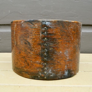 May include: A cylindrical wooden container with a dark, aged appearance. The wood grain is visible, and the container is held together with small metal rivets. The color is a mix of brown and dark tones, suggesting age and use.