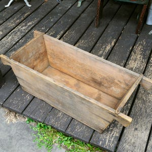 May include: A weathered, rectangular wooden trough with side handles. The trough is made of light brown wood and sits on a dark wooden deck. The trough is empty and appears to be an antique.