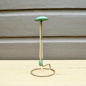 May include: A vintage hat stand with a green top and a gold-colored metal base. The stand has a slender, vertical rod with a coiled section and a decorative bead. The base is a circular ring.