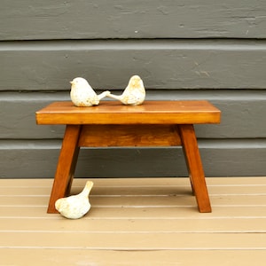 May include: A wooden stool with a natural wood finish. The stool has a simple design with three legs and a rectangular top. Three white ceramic bird figurines are placed on top of the stool.