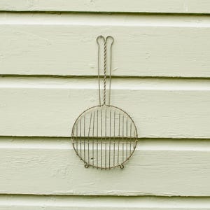 May include: A round, wire mesh grill with two handles hanging on a light green wall.