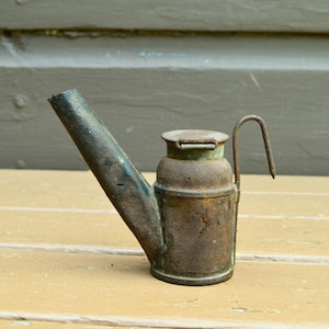 May include: A rusty metal oil lamp with a long spout and a handle. The lamp has a hinged lid and is made of a dark metal.