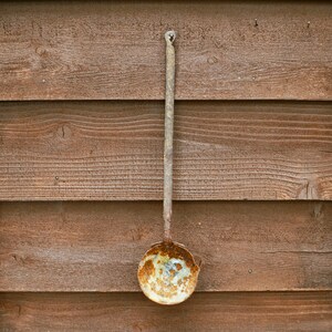 May include: An antique metal ladle with a long handle and a round bowl, showing signs of rust and wear. The ladle is hanging against a wooden plank wall, showcasing its aged appearance and rustic charm.