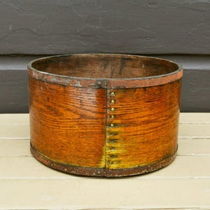 May include: A round, wooden container with a warm, orange-brown hue. The container is reinforced with metal bands and rivets, showing signs of age and use. The wood grain is visible, adding to its rustic charm.